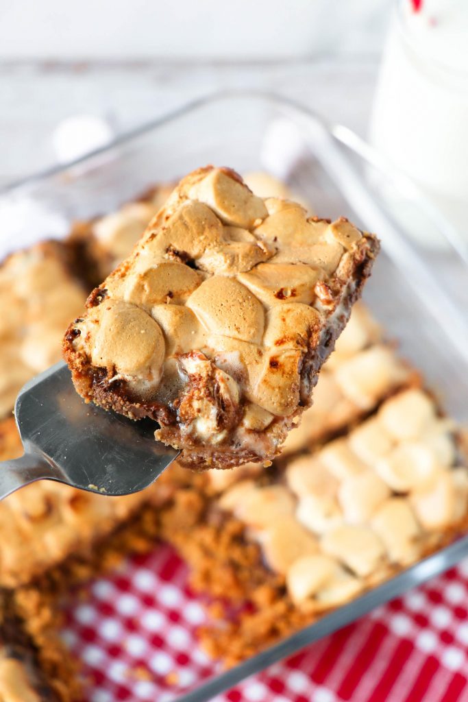 Baking dish of s'mores bars with a piece cut out and held on a serving utensil.