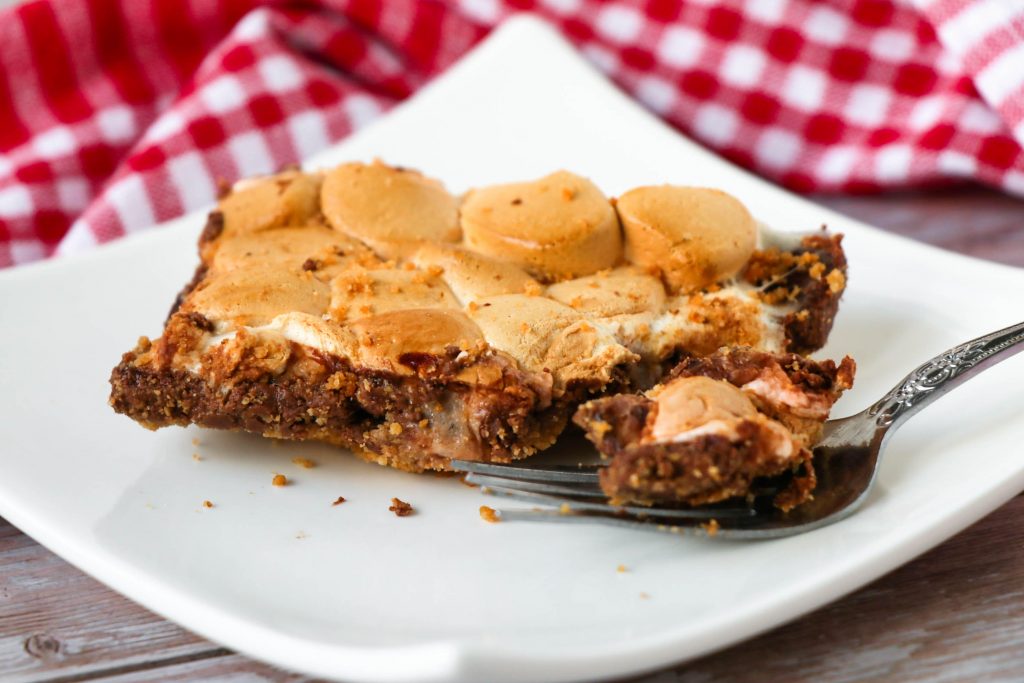 slice of s'mores bars cut and set on a plate with a fork holding a bite of the s'mores bar.